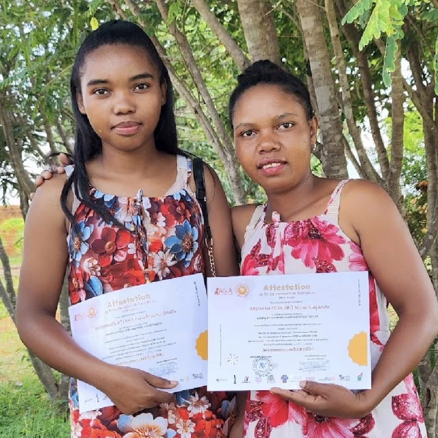 Deux jeunes diplômées en robes fleuries posent avec leurs attestations de formation en couture dans le jardin du Village Aïna