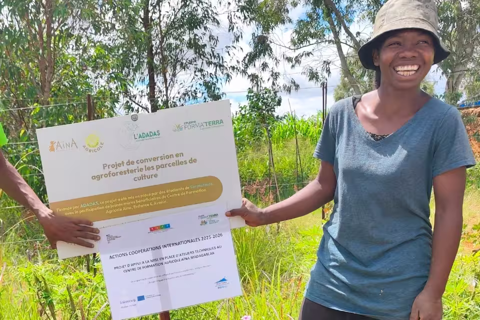 Une maman bénéficiaire sourit en tenant le panneau « Projet de conversion en agroforesterie » porté par ADADAS, Aïna et Formaterra.
