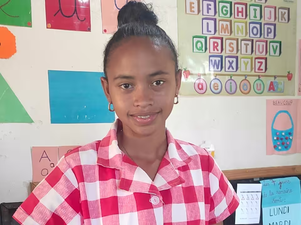Portrait souriant de Mme Sigrie, aide-éducatrice à la maternelle solidaire d'Aïna, en blouse à carreaux rouges, devant le mur pédagogique de sa classe de Moyenne Section.