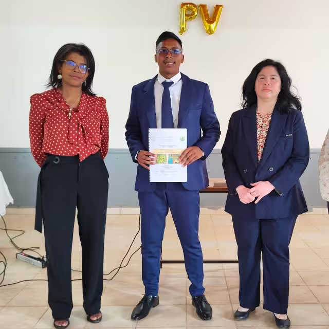 Julien en costume bleu présente son diplôme entre deux membres du jury, sous des ballons dorés « PV », à l'Université MAGIS Madagascar.