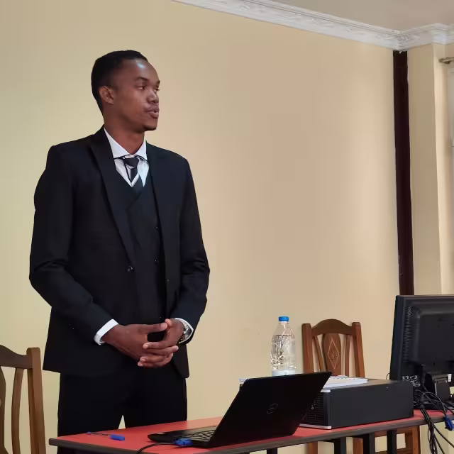 Nantenaina debout en costume sombre, présentant son mémoire d'Audit et Contrôle de Gestion devant l'ordinateur de soutenance.