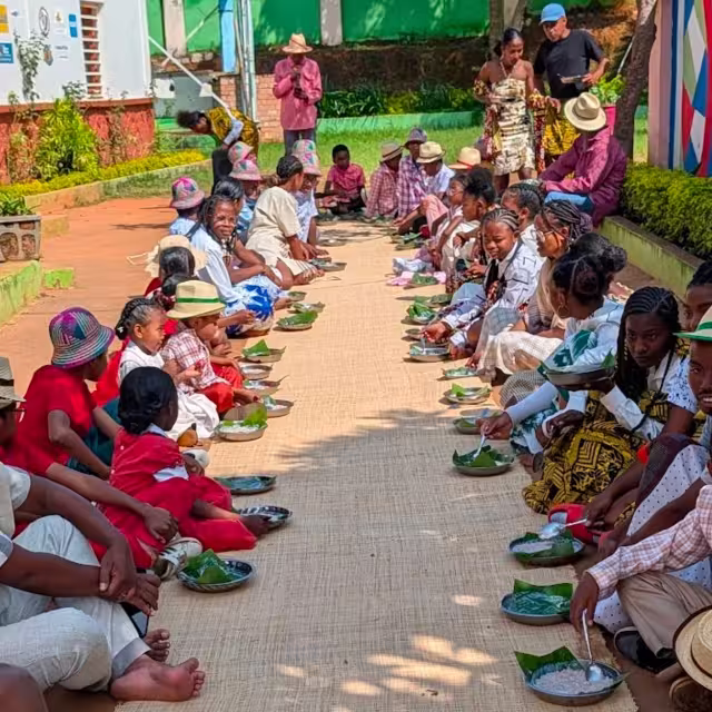 Repas collectif du Taombaovao Malagasy : enfants et jeunes alignés en deux rangées partagent leurs plats servis sur des feuilles de banane.