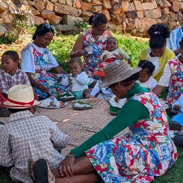 Petit groupe de femmes et de tout-petits partagent le repas du Taombaovao Malagasy à l'extérieur, assis sur une natte au pied d'un mur de pierres.