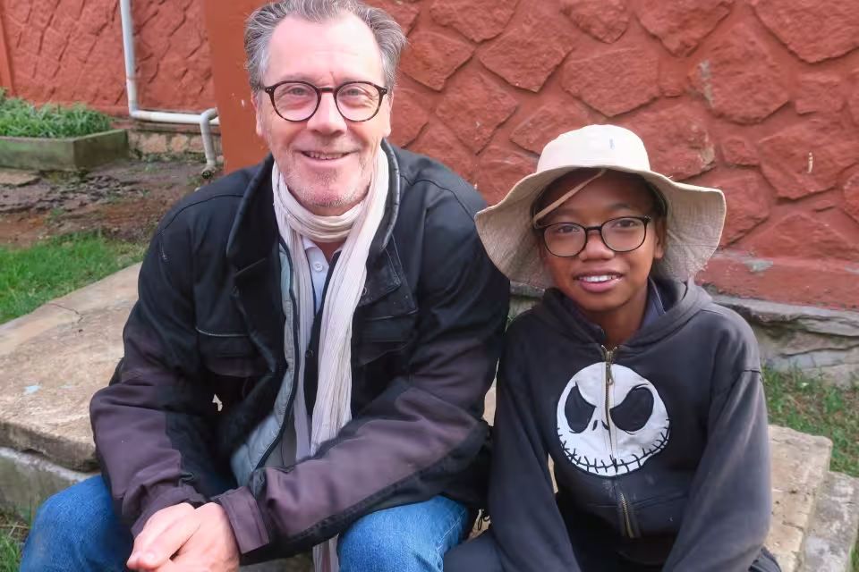 Portrait d'un membre du groupe de motards souriant aux côtés d'un enfant à chapeau et lunettes de l'orphelinat Aïna à Malaza.