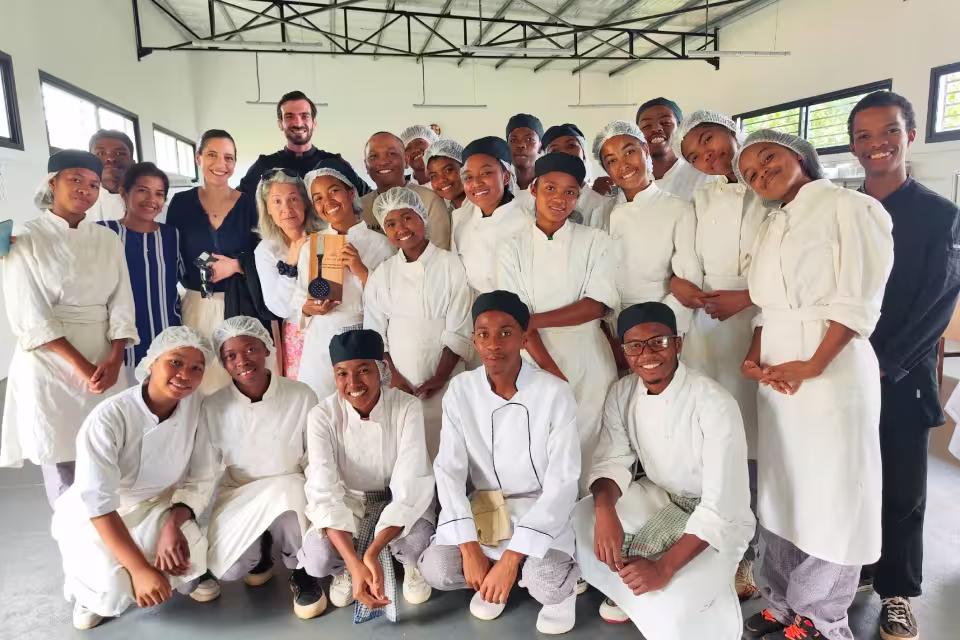 Photo de groupe au sein du laboratoire culinaire avec Laure Kermen, sa collaboratrice et l'ensemble des jeunes apprenants de l'École de Cuisine Solidaire Aïna en tenue de chef.