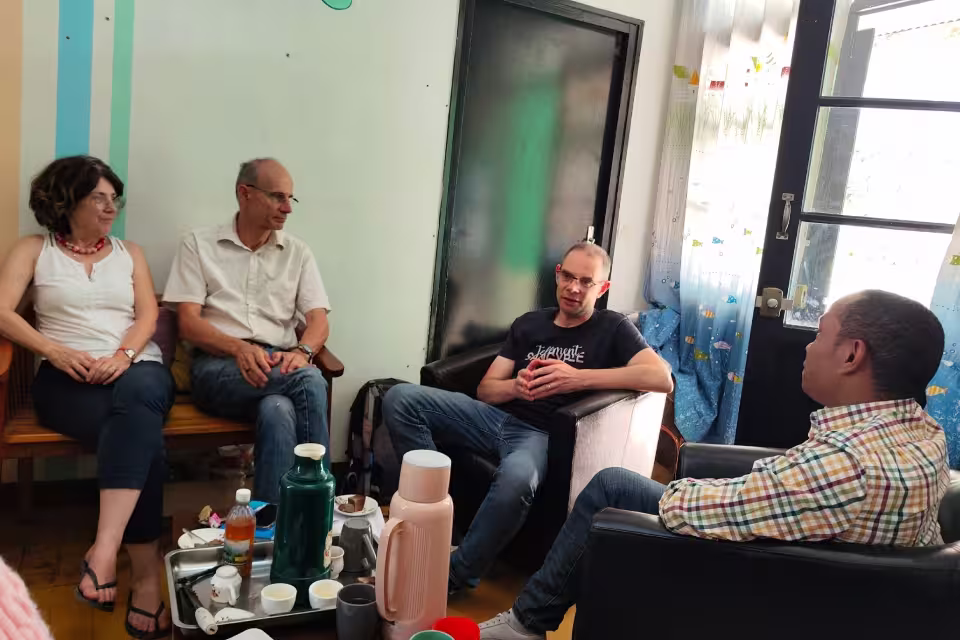 Réunion d'échange en intérieur entre Xavier Lorain, les volontaires Isabelle et Laurent et un responsable d'Aïna autour d'une table chargée de boissons et de gâteaux.