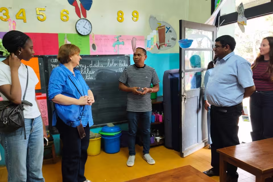 Visiteurs de la Fondation Deloitte en discussion avec un responsable d'Aïna dans une salle de classe maternelle décorée de chiffres et d'affichages pédagogiques en malagasy.