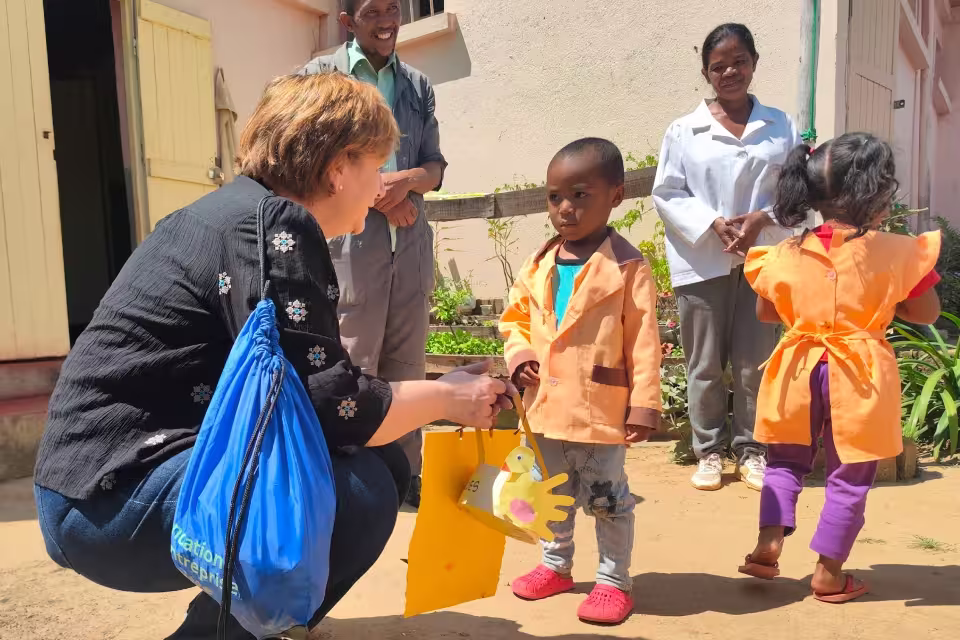 Guilène Bertin, marraine d'un enfant de l'orphelinat de Malaza, accroupie pour offrir un sachet cadeau à son filleul en blouse orange.