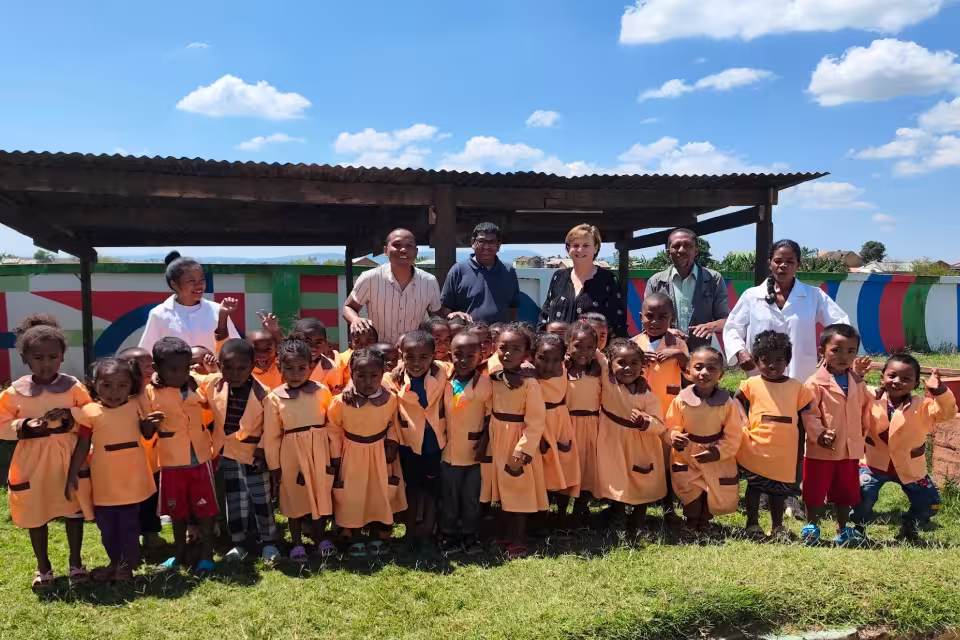 Photo de groupe de la Fondation Deloitte sous un préau avec les enfants en blouse orange et les éducatrices de l'école maternelle solidaire d'Aïna.