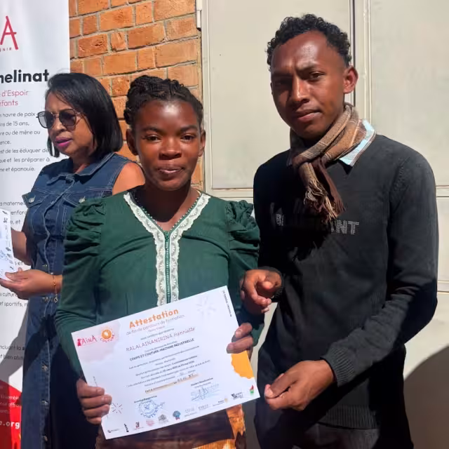 Une diplômée reçoit son attestation CFSC lors de la cérémonie de remise au Village Aïna, devant la bannière Aïna.