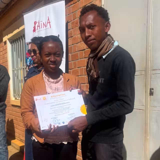 Remise d'attestation CFSC à une diplômée souriante en veste orange lors de la cérémonie de sortie au Village Aïna.
