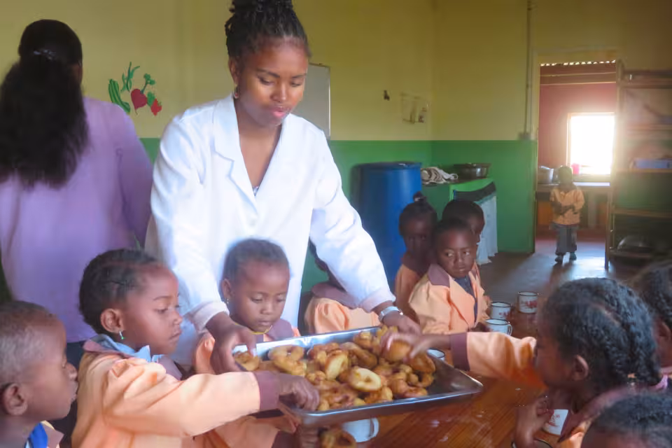 Auxiliaire de cantine en blouse blanche servant un goûter aux enfants de maternelle Aïna en uniforme orange