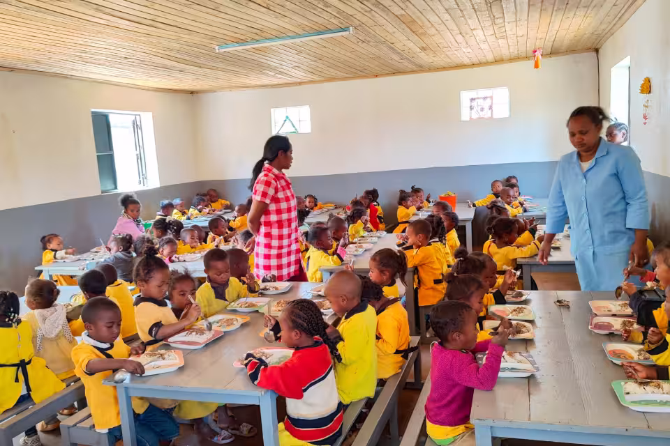 Grande salle de cantine Aïna remplie d'enfants en uniforme jaune déjeunant sous la surveillance de deux éducatrices