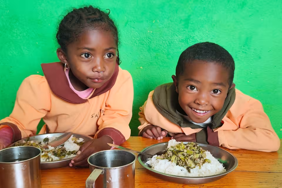 Deux enfants en uniforme orange posant souriants à la table de la cantine Aïna, assiettes de riz et légumes devant eux