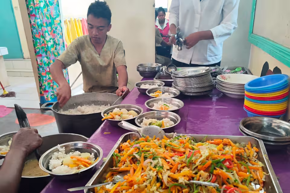 Cuisinier de la cantine Aïna portionnant le riz pour le repas des enfants, légumes colorés en attente sur la table