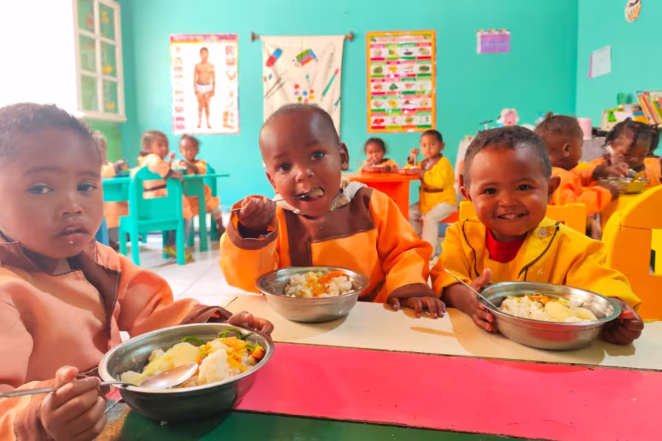 Trois tout-petits de maternelle en tablier orange déjeunant dans leurs bols inox, salle de classe aux murs turquoise