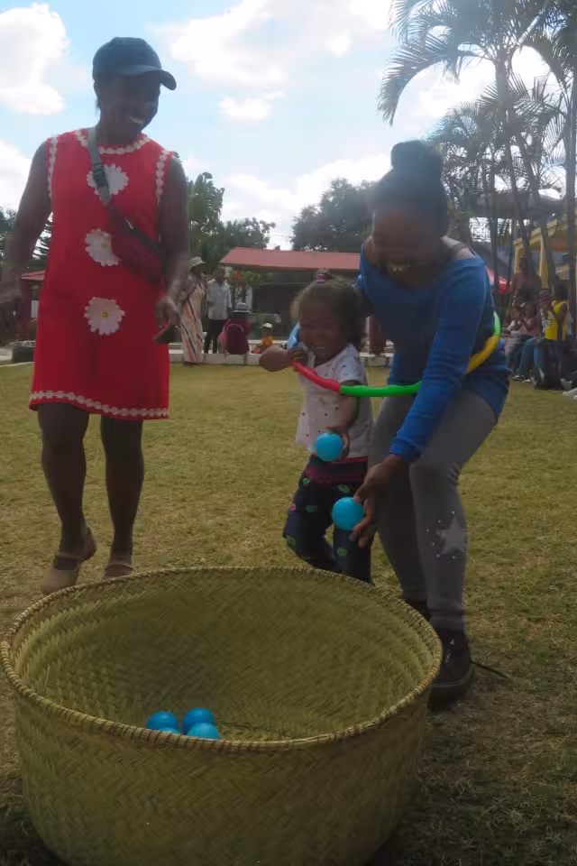 Deux mamans aident une fillette à lancer des balles dans un grand panier tressé lors d'un jeu collectif